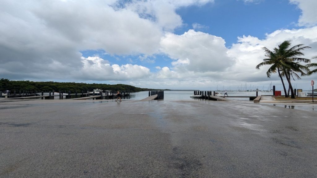 Wide view of a calm South Miami boat ramp with docks, boats, and palm trees under a partly cloudy sky
